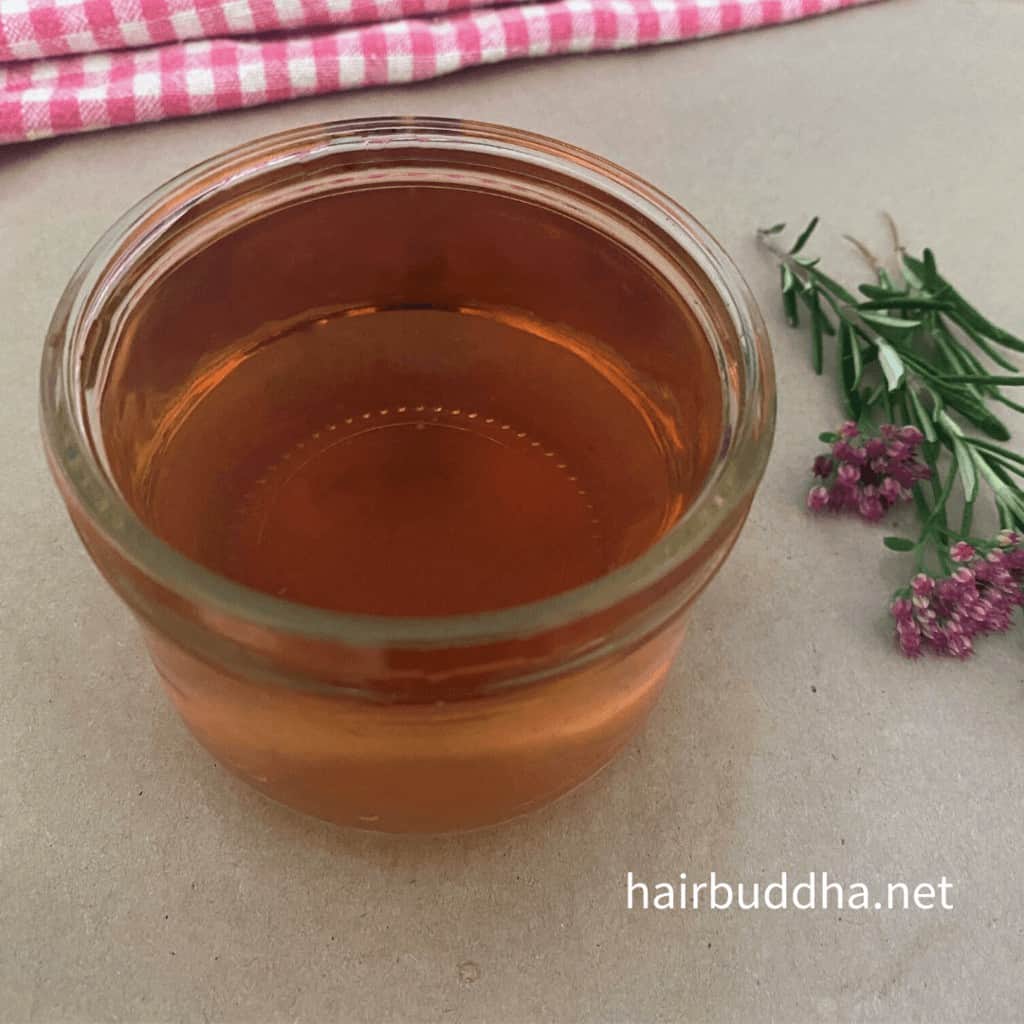 Redish-brown colour Rosemary Water in a glass jar, and on the side are fresh rosemary leaves and red flowers